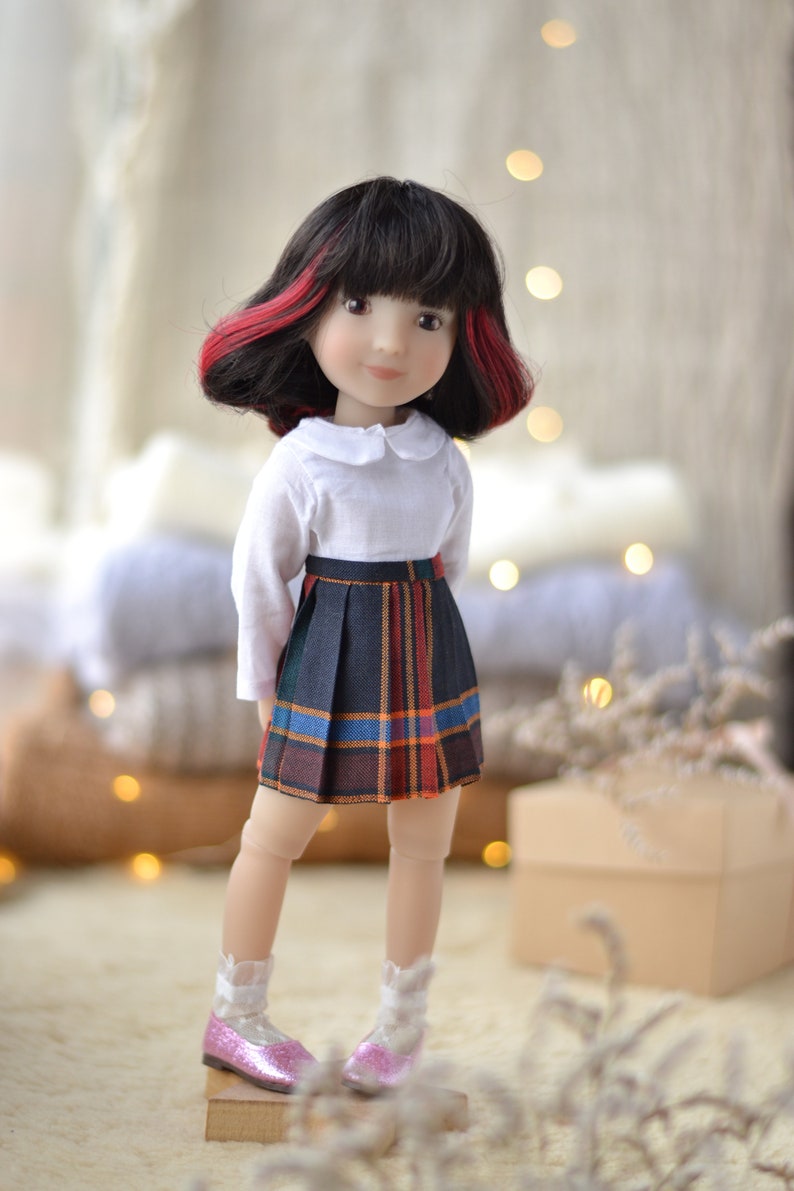 May include: A doll with black hair with red streaks, wearing a white collared shirt and a plaid skirt. The doll is wearing pink shoes and is standing on a wooden platform.