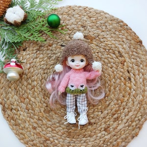 May include: A pink knitted sweater with a green and white sheep design, worn by a doll with pink hair and a brown fuzzy hat. The doll is holding white pom-poms in each hand and is sitting on a woven straw mat.