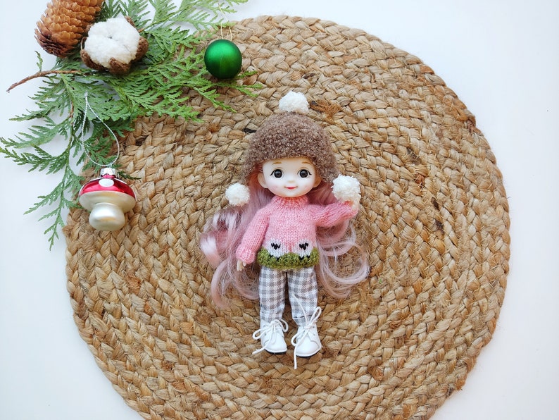 May include: A pink knitted sweater with a green and white sheep design, worn by a doll with pink hair and a brown fuzzy hat. The doll is holding white pom-poms in each hand and is sitting on a woven straw mat.