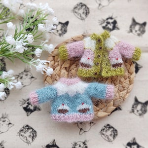 May include: Two knitted cardigans for dolls, one in green and pink with a white trim, and one in blue and pink with a white trim. Both cardigans have a button closure and are decorated with embroidered cupcake designs.