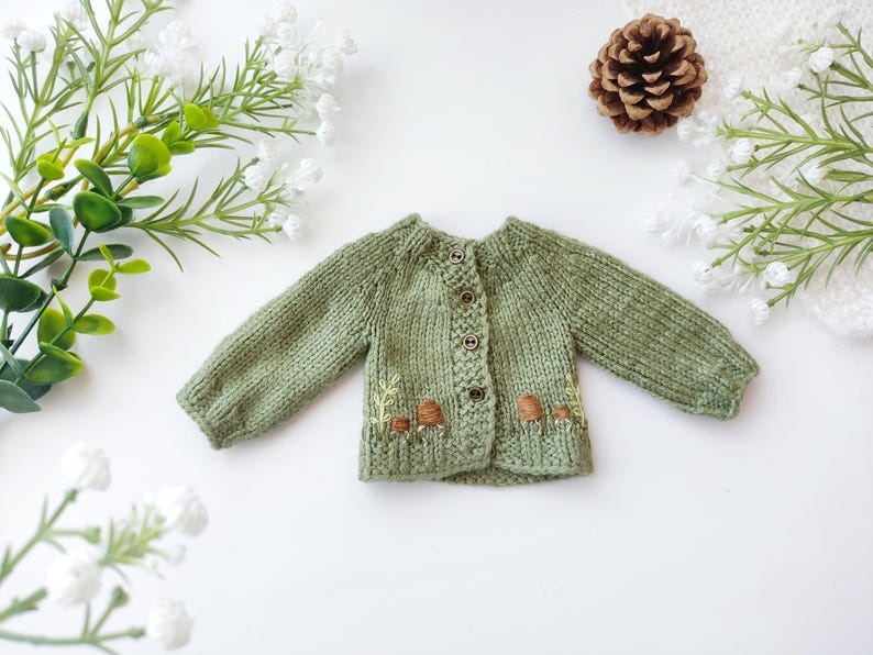 Op de afbeelding: Een saliegroen gebreid vest met lange mouwen en een ronde halslijn. De baby trui heeft geborduurde paddenstoelen en bladeren aan de onderkant. Het vest heeft drie knopen aan de voorkant.