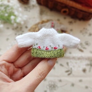 May include: A white knitted sweater with a green and red embroidered trim. The trim features a row of red mushrooms with green stems.