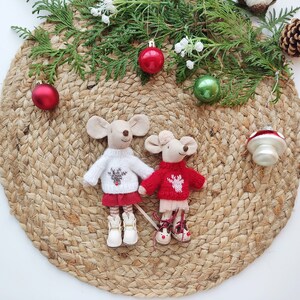 May include: Two small knitted mouse figurines wearing red and white sweaters with reindeer designs. The mice are standing on a woven jute mat with Christmas ornaments and greenery.