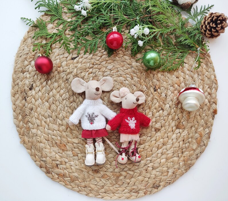 May include: Two small knitted mouse figurines wearing red and white sweaters with reindeer designs. The mice are standing on a woven jute mat with Christmas ornaments and greenery.