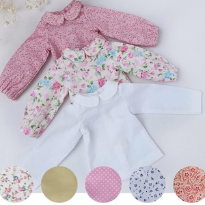 Puede incluir: Tres camisas de manga larga con cuello bebé. Una camisa es de color blanco liso, otra presenta un estampado floral con flores rosas y azules sobre fondo crema, y otra es de tela rosa con un pequeño y denso estampado floral rosa oscuro.
