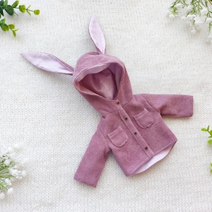 Może przedstawiać: Fioletowa sztruksowa kurtka z kapturem i uszami królika. Kurtka ma dwie przednie kieszenie i jest zapięta na guziki. Tło to biała, teksturowana powierzchnia z gałązkami zieleni i białymi kwiatami.