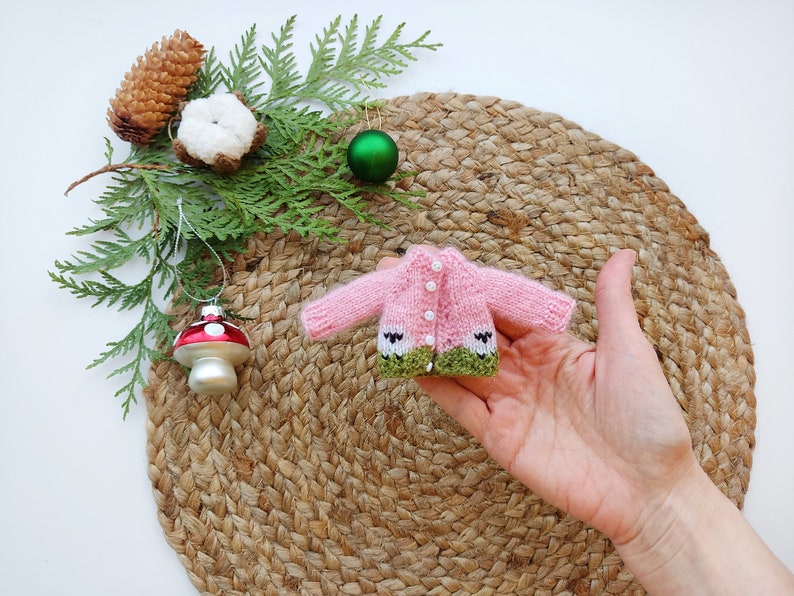 May include: A pink knitted cardigan with green trim and white buttons. The cardigan is small enough to fit in the palm of a hand.