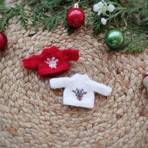 May include: Two miniature knitted sweaters with reindeer designs. The red sweater has a white reindeer with brown antlers. The white sweater has a brown reindeer with a red nose. The sweaters are on a woven straw mat with Christmas ornaments.