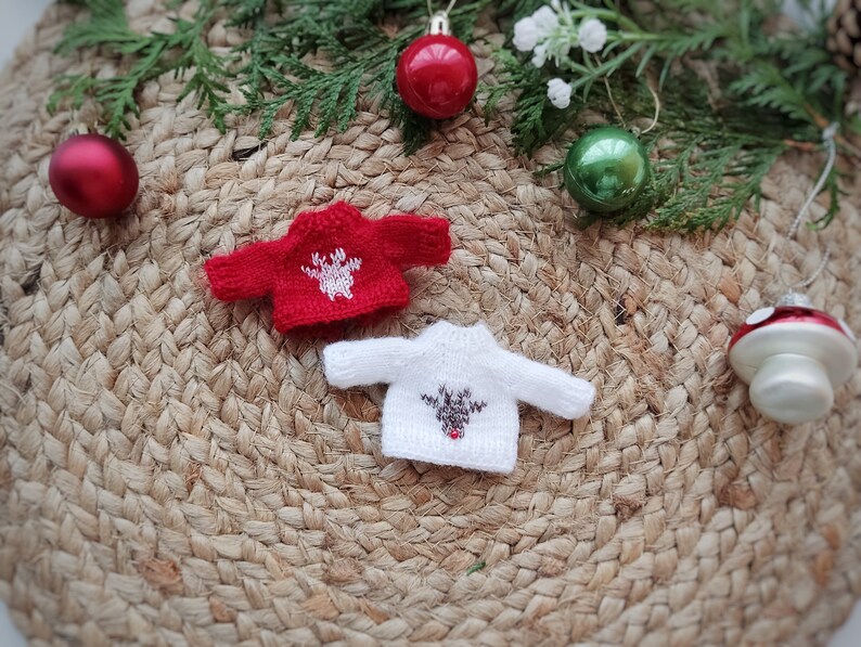 May include: Two miniature knitted sweaters with reindeer designs. The red sweater has a white reindeer with brown antlers. The white sweater has a brown reindeer with a red nose. The sweaters are on a woven straw mat with Christmas ornaments.