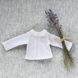 May include: A white long-sleeved shirt with a peter pan collar. The shirt is laid flat on a gray textured surface. A bundle of dried lavender is to the right of the shirt.