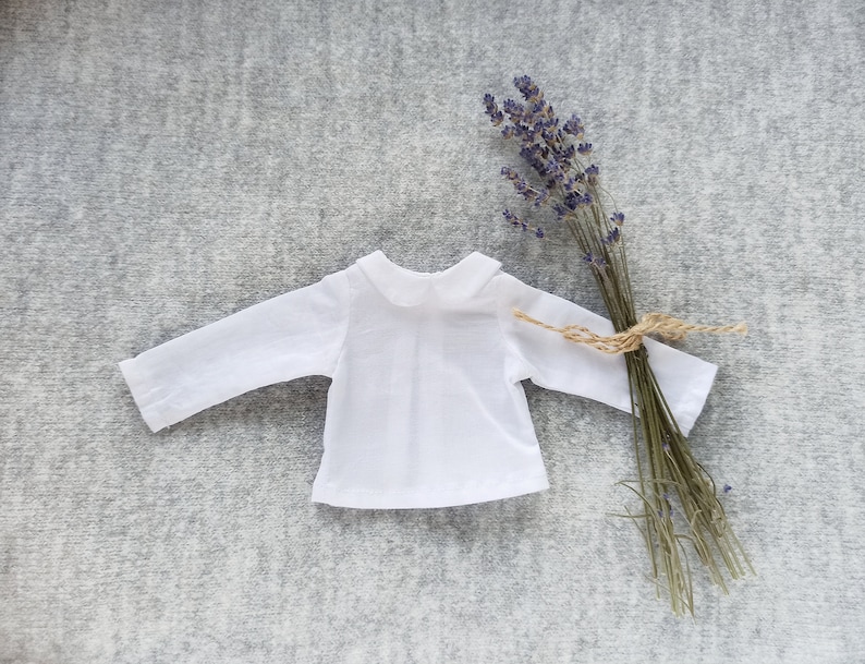 May include: A white long-sleeved shirt with a peter pan collar. The shirt is laid flat on a gray textured surface. A bundle of dried lavender is to the right of the shirt.