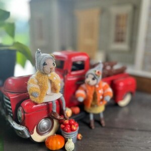 May include: Two small, plush, handmade toys dressed in fall-themed outfits sit in front of a red toy truck. The truck is loaded with miniature pumpkins and gourds. The toys are wearing yellow and orange sweaters and hats.