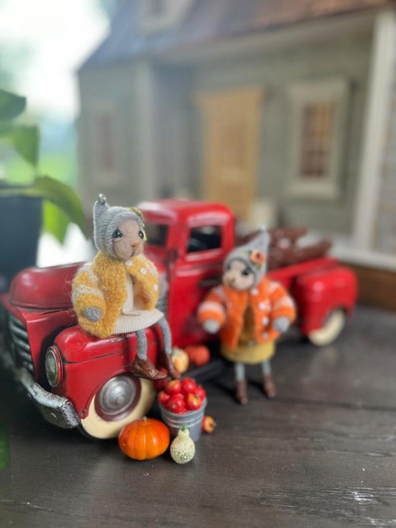 May include: Two small, plush, handmade toys dressed in fall-themed outfits sit in front of a red toy truck. The truck is loaded with miniature pumpkins and gourds. The toys are wearing yellow and orange sweaters and hats.