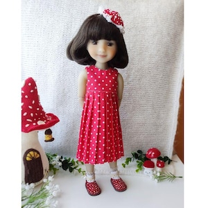 Puede incluir: Una muñeca con un vestido rojo de lunares blancos, zapatos a juego y una diadema. La muñeca tiene el pelo corto y oscuro y está de pie frente a un fondo blanco. También hay una casa de setas y pequeñas decoraciones de setas.