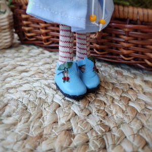 May include: A pair of blue doll shoes with red berry accents and green leaves. The shoes are on a woven straw mat.