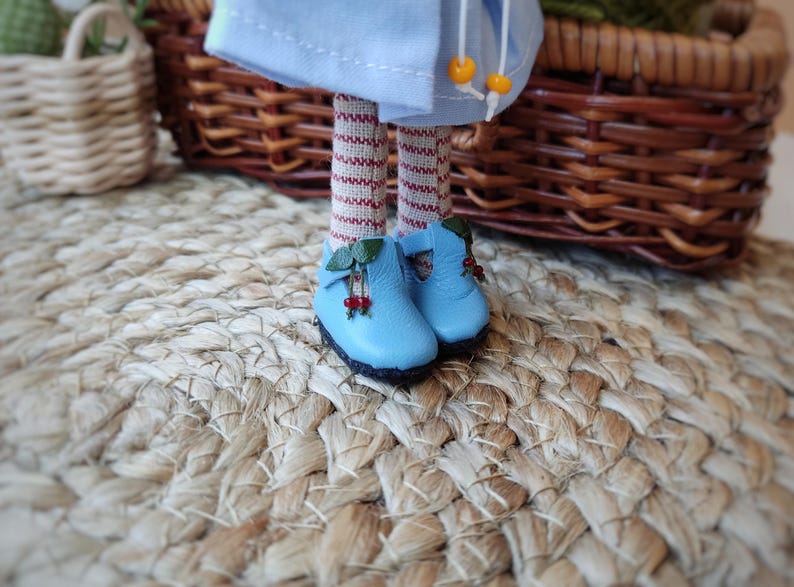 May include: A pair of blue doll shoes with red berry accents and green leaves. The shoes are on a woven straw mat.