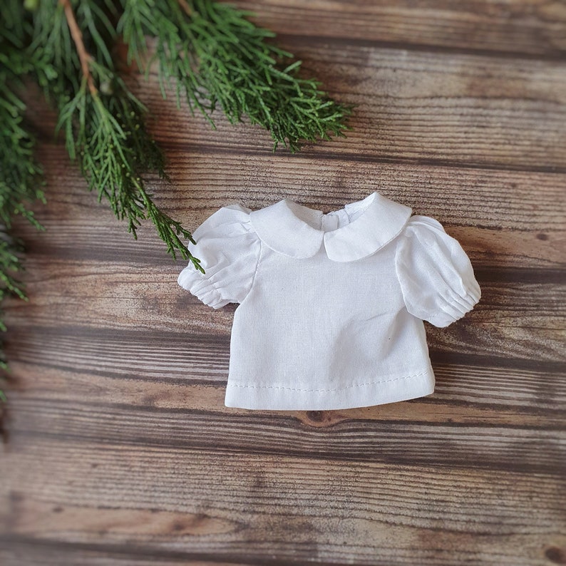 May include: A white cotton doll shirt with a peter pan collar and puffed sleeves.