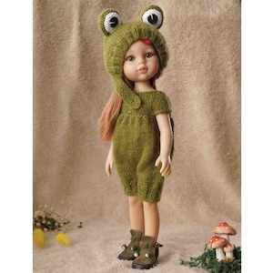 Może przedstawiać: Lalka ubrana w zielony, dziergany kostium żaby, w tym czapkę z dużymi oczami i kombinezon. Lalka ma długie, jasnobrązowe włosy i zielone buty z żabimi detalami. Mała dekoracja z grzybami znajduje się w tle.