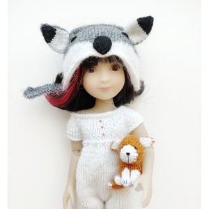 May include: A doll wearing a gray and white knitted wolf hat with black ears and a white knitted jumpsuit. The doll is holding a small knitted orange and white puppy.