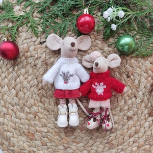 May include: Two plush mouse toys wearing red and white sweaters with reindeer designs. The mice are standing on a woven brown mat with green pine branches and red and green Christmas ornaments.