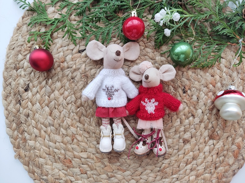 May include: Two plush mouse toys wearing red and white sweaters with reindeer designs. The mice are standing on a woven brown mat with green pine branches and red and green Christmas ornaments.