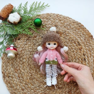 May include: A pink and green knitted sweater with a brown fur hat and white pom poms on a doll with pink hair. The doll is sitting on a woven straw mat.