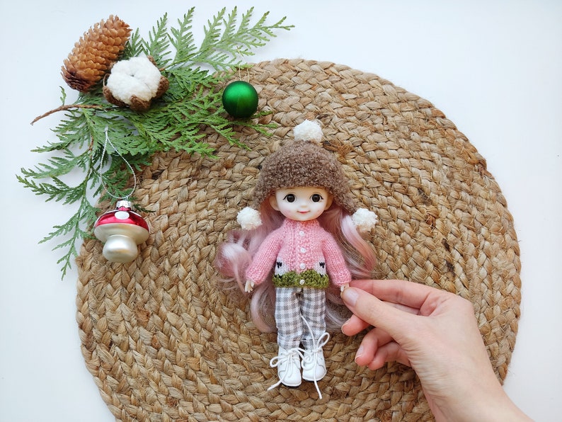 May include: A pink and green knitted sweater with a brown fur hat and white pom poms on a doll with pink hair. The doll is sitting on a woven straw mat.