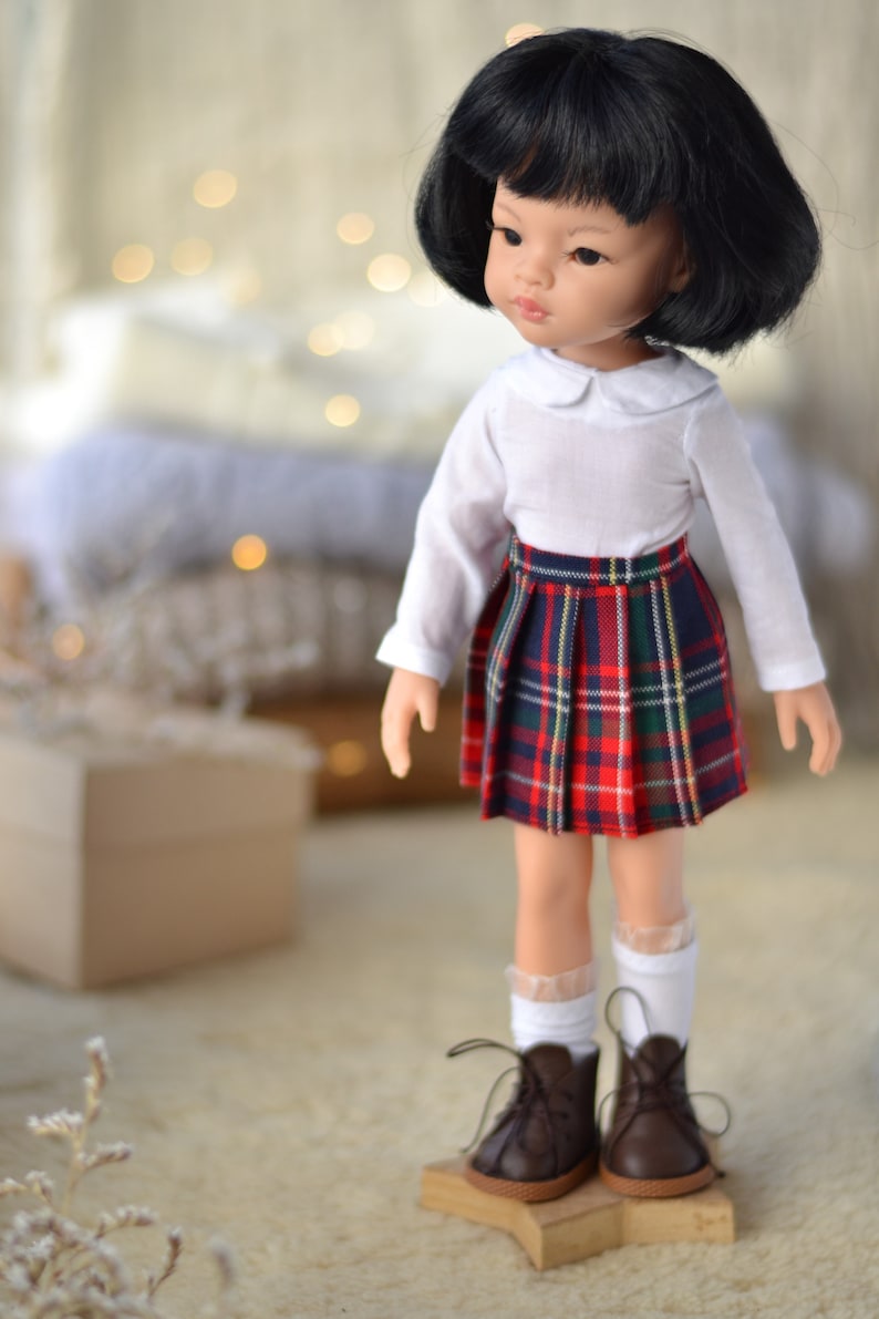 May include: A doll with black hair wearing a white collared shirt and a red and black plaid skirt. The doll is wearing brown shoes and white socks. The doll is standing on a wooden platform.