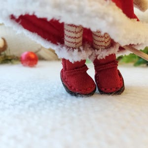 Könnte beinhalten: Rote Filzstiefel mit weißem Besatz und einem rot-weiß gestreiften Strumpf. Die Stiefel stehen auf einer weißen, flauschigen Oberfläche.