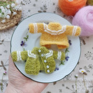 May include: Two miniature knitted sweaters, one yellow with white trim and one green with white trim, on a white plate with floral designs.