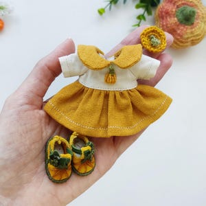 May include: A miniature doll outfit featuring a mustard yellow dress with a white top and collar, a matching hair accessory, and tiny yellow shoes with green accents. The outfit is designed with a pumpkin theme.