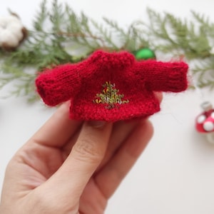 Op de afbeelding: Een klein rood gebreid truitje met een geborduurd kerstboomontwerp in groen en goud.