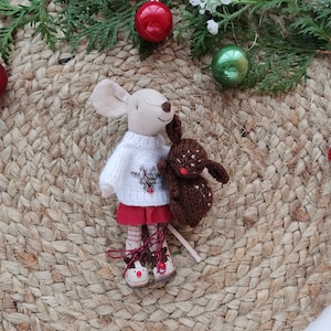 May include: A plush toy mouse wearing a white sweater with a reindeer design and a red skirt, holding a brown plush toy bunny. The toys are on a woven jute mat with red and green Christmas ornaments.