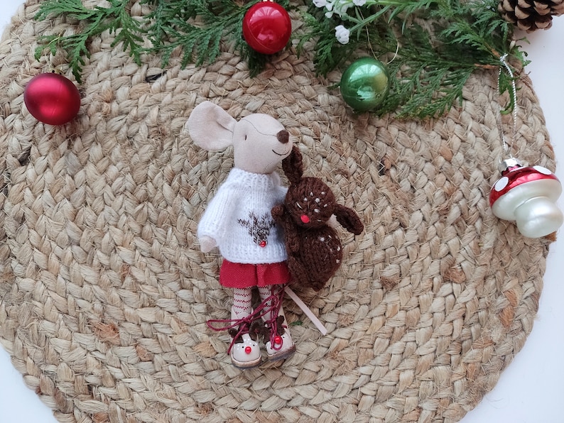 May include: A plush toy mouse wearing a white sweater with a reindeer design and a red skirt, holding a brown plush toy bunny. The toys are on a woven jute mat with red and green Christmas ornaments.
