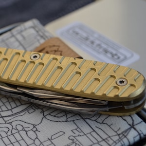 Könnte beinhalten: Ein goldenes Multitool mit einem strukturierten Griff und mehreren Klingen. Das Werkzeug liegt auf einer gemusterten Stoffoberfläche.