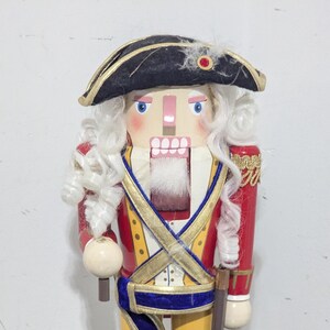 May include: A wooden nutcracker soldier in a red coat, yellow pants, and black boots. The nutcracker has a white wig, a black hat, and a detailed face. It stands on a green base.