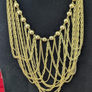 Puede incluir: Un collar dorado con múltiples hilos de diferentes estilos de cadena. El collar presenta una serie de pequeñas cuentas redondas a lo largo del escote. Las cadenas caen en cascada hacia abajo, creando un efecto de capas. La joya se exhibe sobre un maniquí negro.