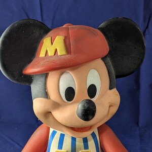May include: A vintage Mickey Mouse figurine wearing a red baseball cap with a yellow "M", a blue and white striped shirt, and blue trousers. The figurine has black ears and a smiling expression.