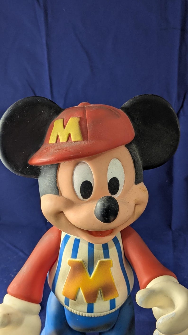 May include: A vintage Mickey Mouse figurine wearing a red baseball cap with a yellow "M", a blue and white striped shirt, and blue trousers. The figurine has black ears and a smiling expression.