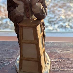 May include: A ceramic figurine of a clown wearing a brown hat and jacket, resting their chin on their hands while leaning over a tall, rectangular, tan and white stool. The clown has a painted face with red accents and is standing on a base.