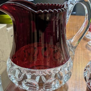 May include: A ruby red glass jug with a clear glass handle. The top rim has a scalloped edge. The bottom portion of the jug has a clear, patterned design. The jug is likely for serving beverages.