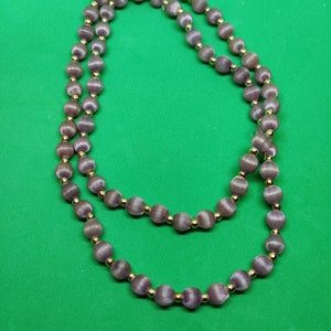 Puede incluir: Un collar largo hecho de cuentas de seda púrpura con detalles dorados. Las cuentas son redondas y tienen un brillo sutil.