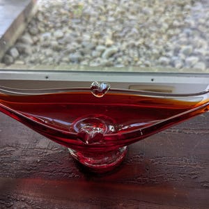 以下が含まれることがあります： ユニークな非対称デザインのヴィンテージルビーレッドガラスボウル。ボウルは、幅広のオープンセンターと優雅に湾曲したエッジを備え、各端には装飾的な透明ガラスのアクセントが付いています。ボウルの長さは約30cmです。