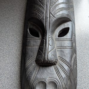 May include: A dark brown, carved wooden mask with a stylized face. The mask features open eye sockets, a prominent nose, and detailed carvings of swirling patterns and lines. The mask is approximately 12 inches long.
