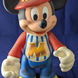 May include: A vintage Mickey Mouse figurine wearing a red cap with a yellow "M", a striped shirt with a yellow "M", blue trousers, and yellow shoes. The figure has black ears and white gloves.