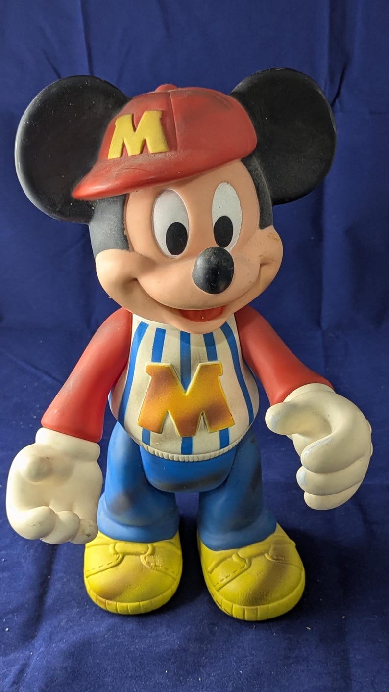 May include: A vintage Mickey Mouse figurine wearing a red cap with a yellow "M", a striped shirt with a yellow "M", blue trousers, and yellow shoes. The figure has black ears and white gloves.
