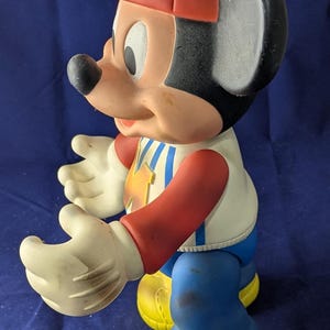 May include: A vintage Mickey Mouse figurine, with a red hat, white gloves, and yellow shoes. The figure is wearing a white shirt with a blue and yellow design, blue trousers, and red sleeves. The classic cartoon character is in a standing pose.