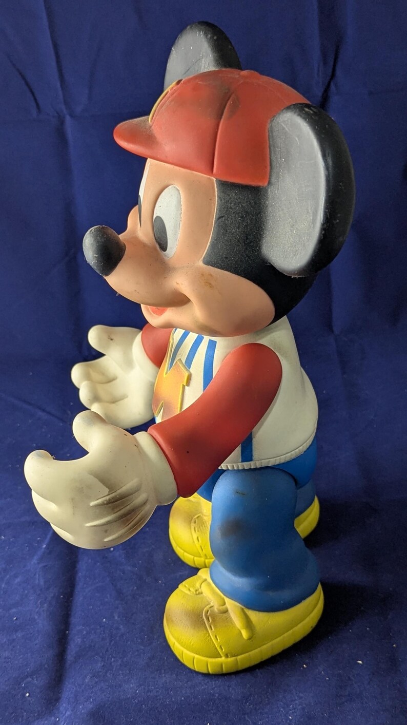 May include: A vintage Mickey Mouse figurine, with a red hat, white gloves, and yellow shoes. The figure is wearing a white shirt with a blue and yellow design, blue trousers, and red sleeves. The classic cartoon character is in a standing pose.