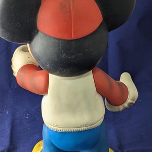 May include: A vintage Mickey Mouse figurine, seen from behind, wearing a red and black cap, white shirt, blue trousers, and yellow shoes. The figure has large black ears and white gloves. The figurine is a collectible item.
