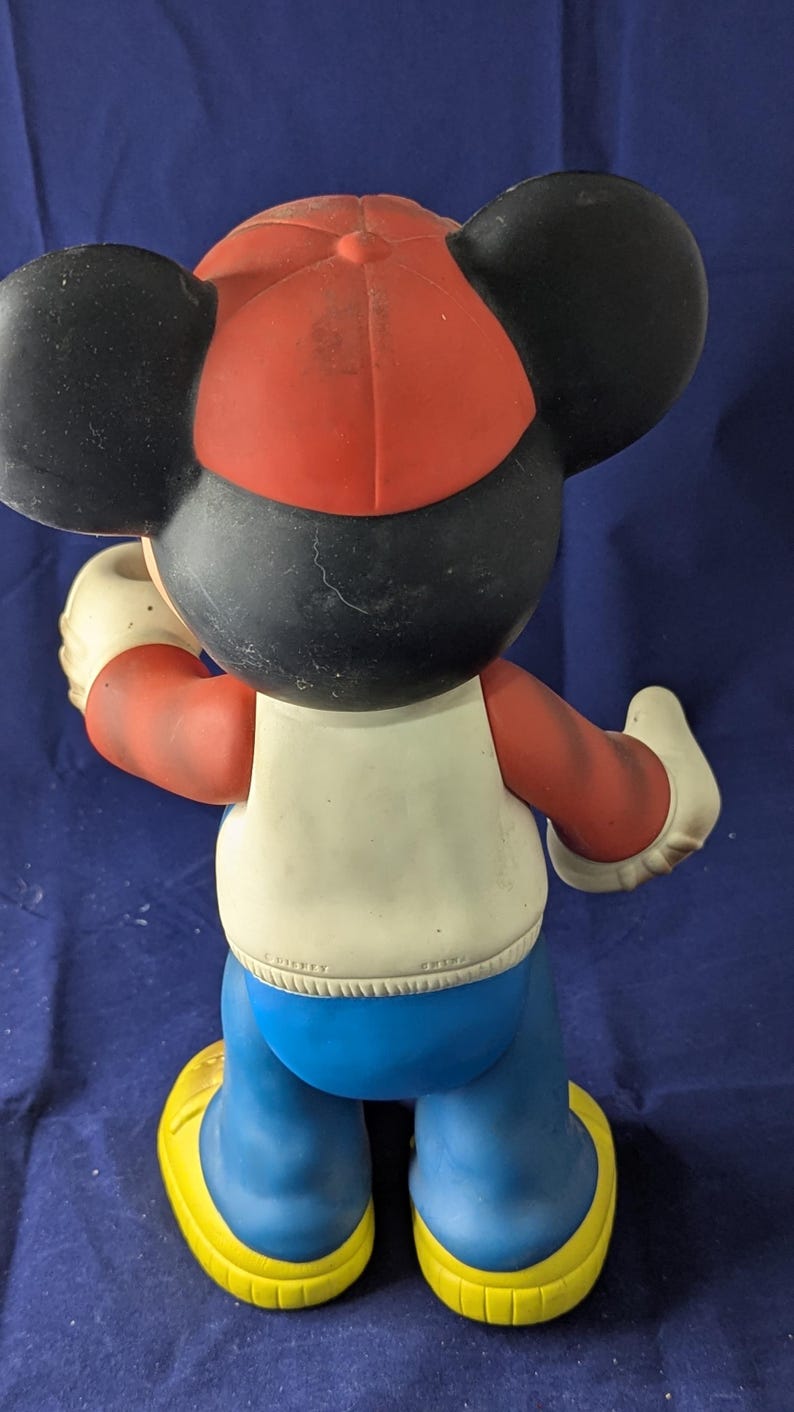 May include: A vintage Mickey Mouse figurine, seen from behind, wearing a red and black cap, white shirt, blue trousers, and yellow shoes. The figure has large black ears and white gloves. The figurine is a collectible item.